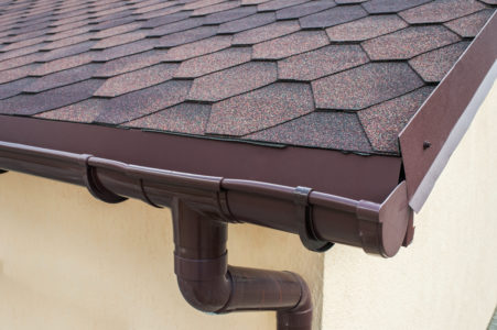The corner of a home's brown roof with brown gutters connected to downspout.