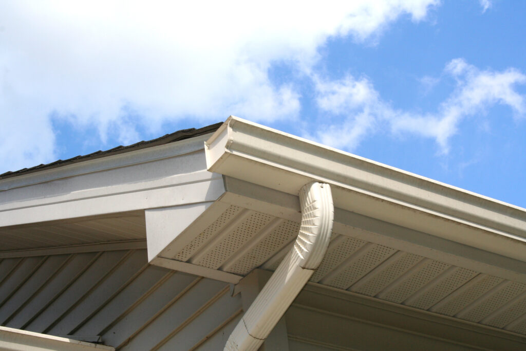 Gutter and downspout of a house with a blue sky and white clouds in the background.